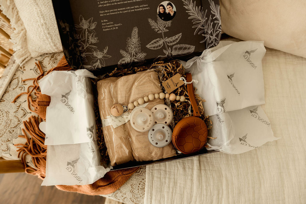 Ninni Co's Cinnamon Sugar baby gift basket is beautifully displayed in a decorative box. It contains a pacifier clip, a pacifier case, a soft baby blanket and three pacifiers.
